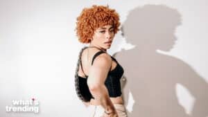 Person with curly orange hair wearing a black crop top and white pants posing against a white wall with a shadow behind them