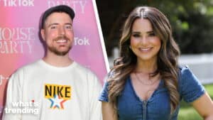 Man in white Nike sweatshirt and black cap smiling next to woman in blue zip-up dress outdoors with white fence background
