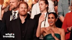 Prince Harry smiling and Meghan Markle waving and smiling at an event with a crowd behind them.
