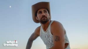 Man wearing a cowboy hat, pearl necklace, and white lace tank top against a clear sky background