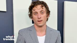 Young man with curly hair wearing a gray checkered blazer and white shirt against a blue and white background
