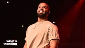 Drake smiling on stage wearing a beige OVO t-shirt and gold necklace with a dark background and red lighting.
