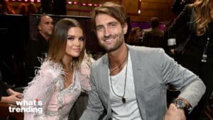 Two people smiling at an event, the woman in a pink feathered dress and the man in a gray blazer with a watch and necklace.