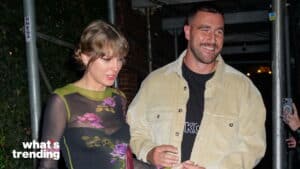 Taylor Swift in a floral sheer top and Travis Kelce in a beige jacket smiling while walking outdoors at night
