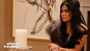 Woman with long black hair wearing a black dress and fur stole sitting indoors with a serious expression