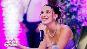 Millie Bobby Brown speaking into a microphone wearing silver jewelry and a purple dress with a blurred purple background