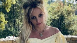 Britney Spears wearing a yellow off-shoulder top and layered necklaces outdoors with trees in the background