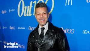 Man in black leather jacket and tie smiling at Out100 event with blue branded backdrop