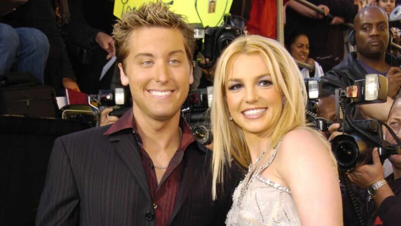 Lance Bass and Britney Spears smiling at a red carpet event with photographers in the background