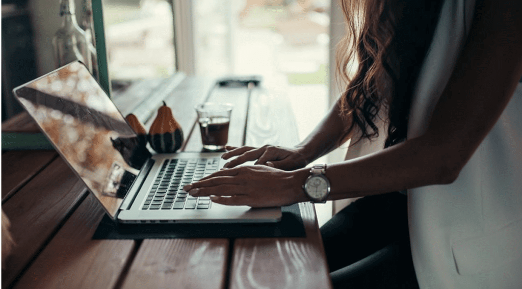 Person typing on a laptop at a wooden table with a glass of coffee and decorative gourds nearby