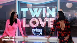 Two women smiling at each other beside a neon sign reading "TWIN LOVE prime" with a city view background