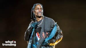 Travis Scott performing on stage wearing a graphic t-shirt, leather jacket, and gloves against a dark background