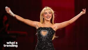 Sabrina Carpenter smiling with arms outstretched, wearing a black dress with a heart-shaped cutout, holding a microphone on stage.