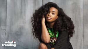 Woman with voluminous curly hair wearing a black graphic t-shirt sitting against a gray backdrop with "what's trending" text
