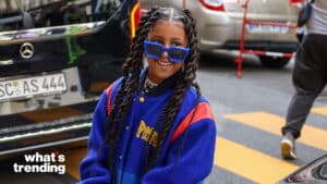 Smiling woman with long braided hair wearing blue sunglasses and a blue jacket with red stripes near a Mercedes car
