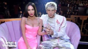 Megan Fox in a pink dress and Machine Gun Kelly in a white suit sitting on a purple couch at an event.