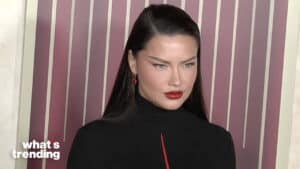 Adriana Lima wearing a black outfit with red lipstick and earrings against a striped background