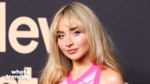 Blonde woman with blue eyes wearing a pink and white patterned top against a dark background with partial text.