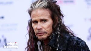 Steven Tyler with long hair and a black jacket at a public event with a blurred background.