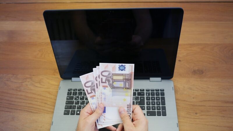 Hands holding multiple 50 euro banknotes in front of a laptop with a black screen on a wooden table.