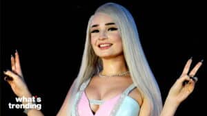 Woman with long platinum blonde hair smiling and making peace signs with both hands against a black background
