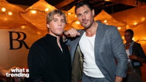 Two men posing indoors under warm lighting, one in a black shirt and the other in a gray suit with a white shirt.