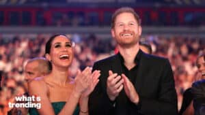 Prince Harry and Meghan Markle smiling and clapping at an event with a blurred crowd background