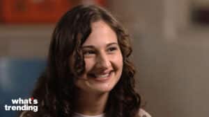 Smiling young woman with curly brown hair in an indoor setting with "what's trending" logo in the corner
