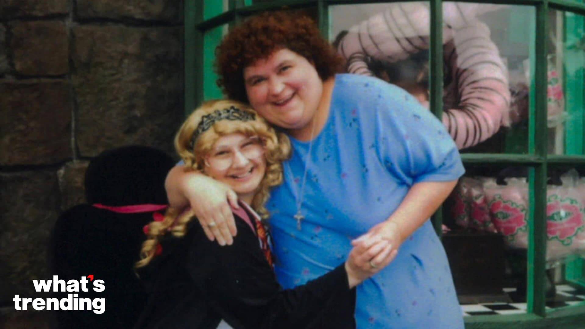 Two smiling women hugging, one with curly hair in a blue shirt and the other with blonde hair and glasses in a black outfit