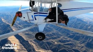 Person hanging out of a small white and blue airplane flying over mountainous terrain