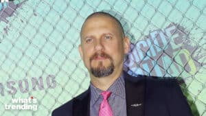 Man with goatee wearing a pink tie and dark suit standing in front of a chain-link fence with colorful graffiti text