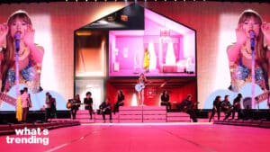 Taylor Swift performing on stage with musicians and a large screen showing a house set and her image