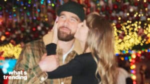 Woman kissing man wearing a beanie and plaid jacket with festive holiday lights in the background