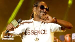 Snoop Dogg smiling and holding a microphone wearing an "ESSENCE" shirt and sunglasses on stage