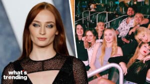 Sophie Turner in a black dress with a sheer top next to a group of smiling people seated in a stadium or arena.