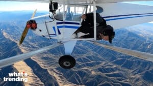 Person hanging out of a small white airplane flying over mountainous terrain with "what's trending" text in corner