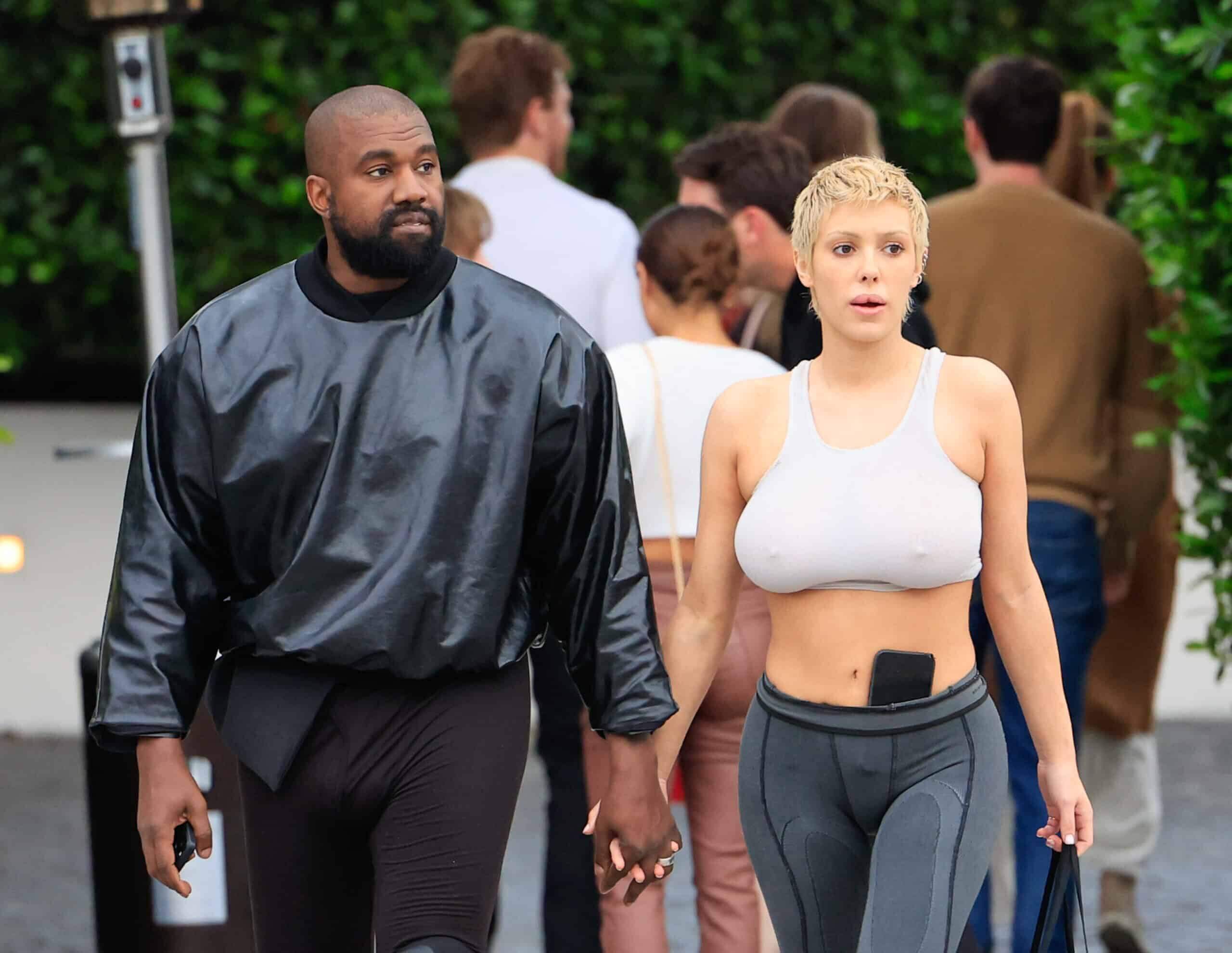 Kanye West and Julia Fox holding hands walking outdoors, Kanye in black oversized top, Julia in white crop top and leggings.