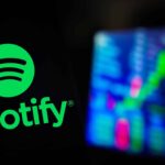 In this photo illustration, the Spotify logo is displayed on a smartphone screen, with graphic representation of the stock market in the background.