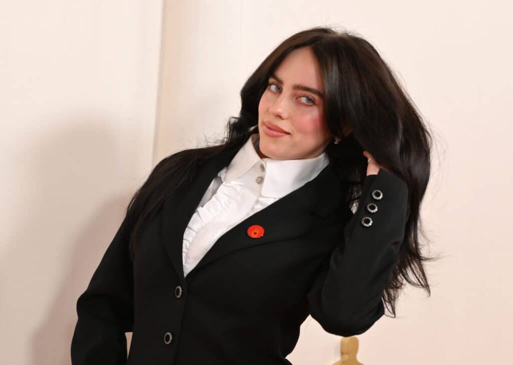 Woman with long dark hair wearing a black blazer and white ruffled shirt, touching her hair and smiling slightly