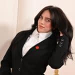 Woman with long dark hair wearing a black blazer and white ruffled shirt, touching her hair and smiling slightly