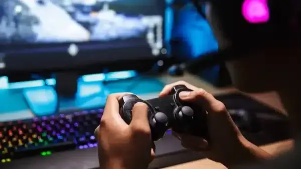 Person holding a black PlayStation controller playing a video game on a PC with a colorful keyboard.