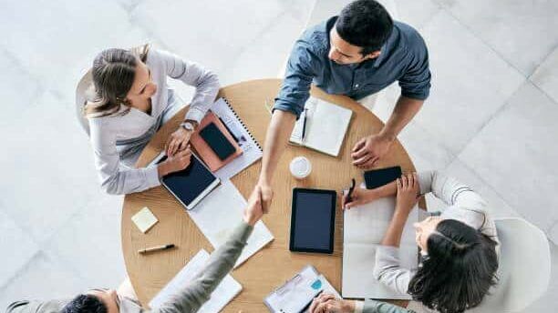 Four colleagues sitting around a table with tablets and notebooks, two shaking hands during a meeting.