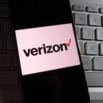 In this photo illustration, Verizon logo is displayed on a smartphone with a workplace background.