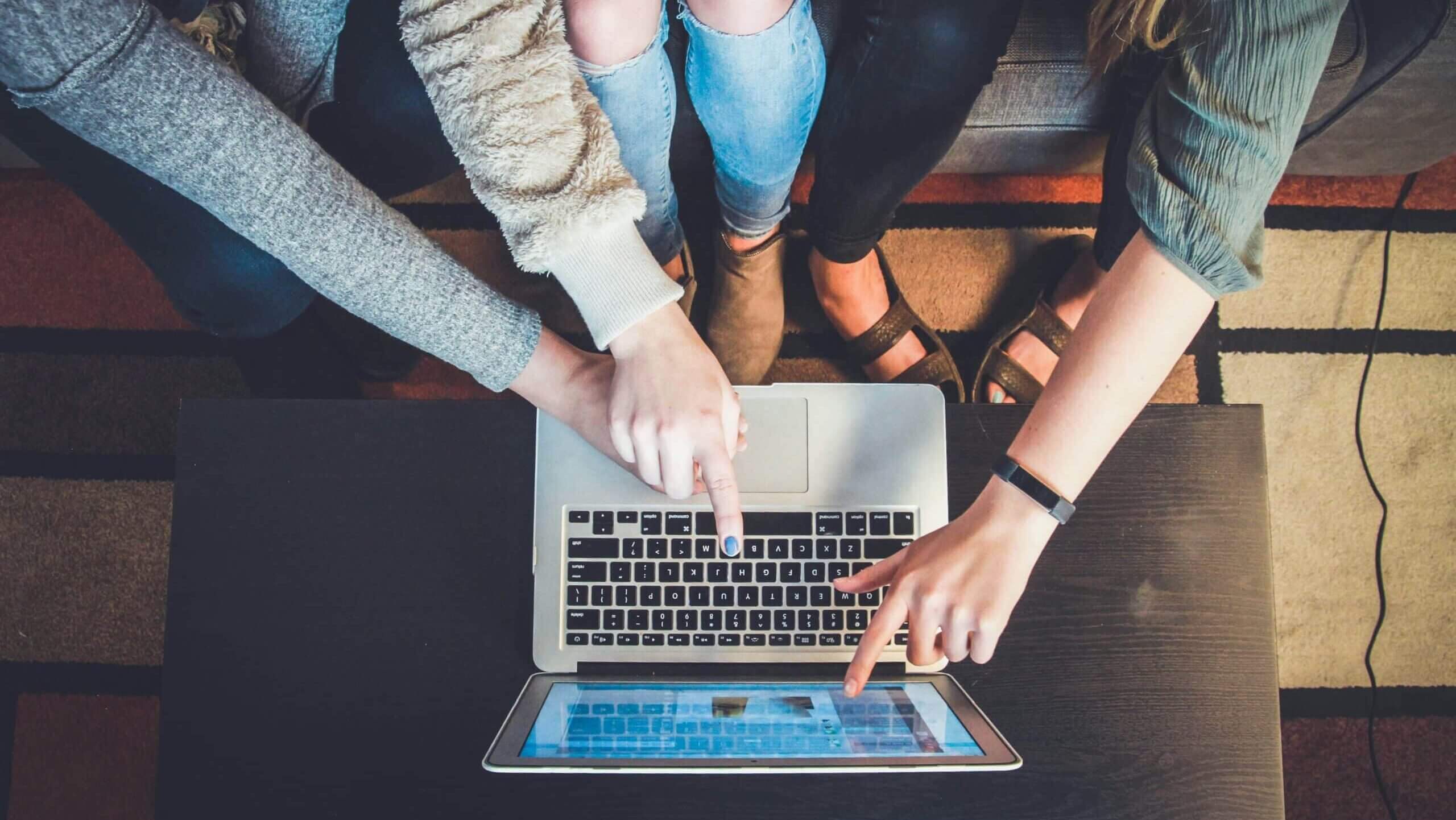 Three people pointing at a laptop screen while sitting on a couch with a patterned rug underneath.