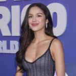 Olivia Rodrigo smiling in a black lace dress at a promotional event with a purple backdrop.