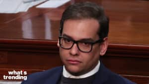Man wearing black glasses and a navy suit sitting in a wooden-paneled room with "what's trending" text in the corner