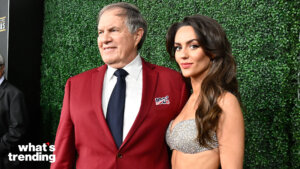 Bill Belichick in a red NFL 100 blazer standing next to a woman in a sparkling silver crop top against a green hedge wall