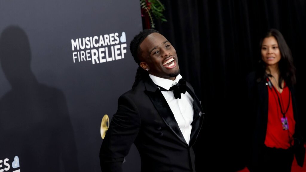 Man in black tuxedo smiling at MusiCares Fire Relief event with woman in red top in background