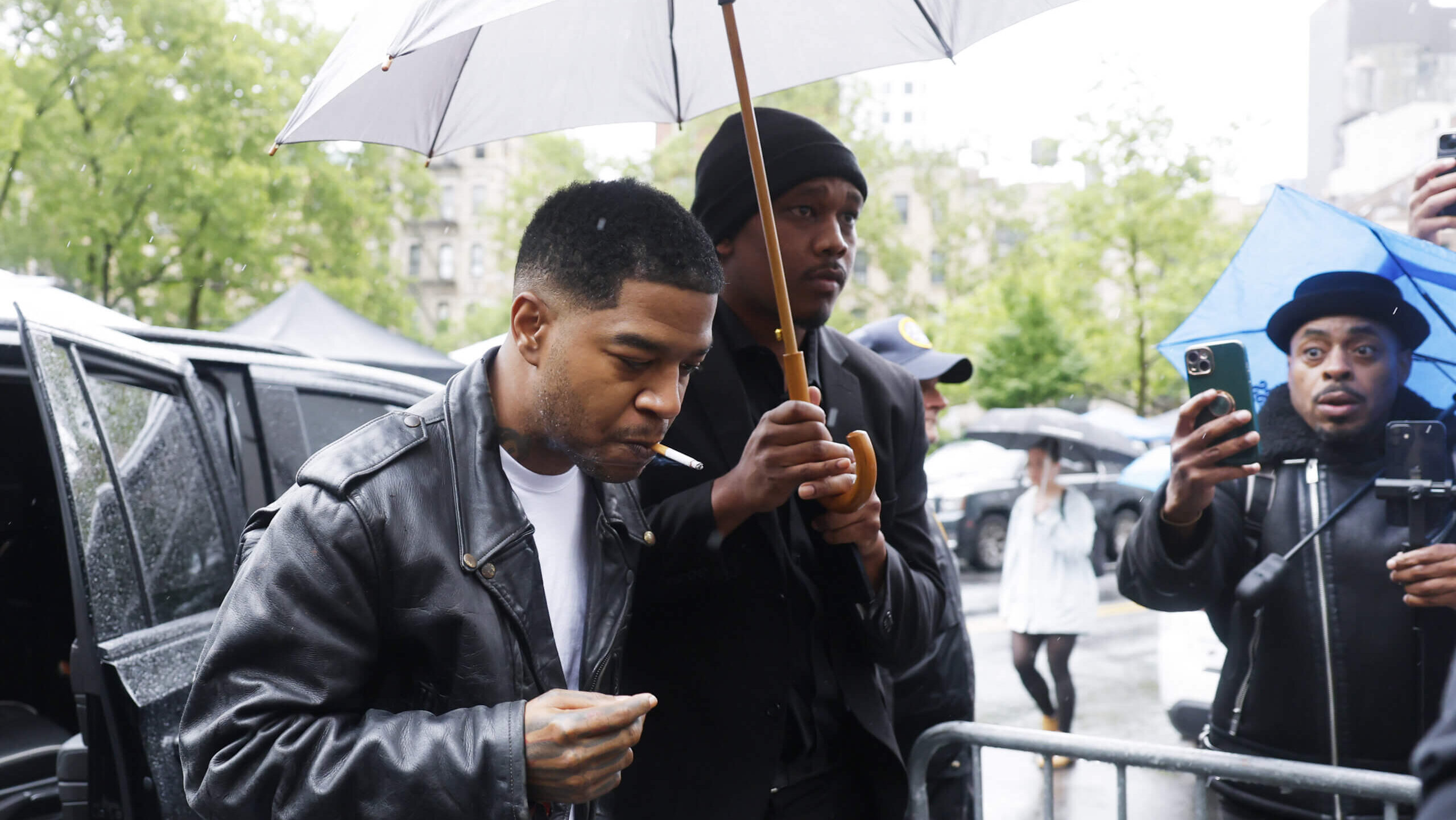 A man in a black leather jacket smoking a cigarette under an umbrella held by another man in black during a rainy day.