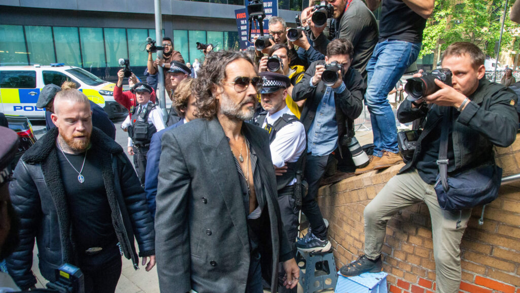 Russell Brand wearing sunglasses and a dark blazer walking past photographers and police officers.